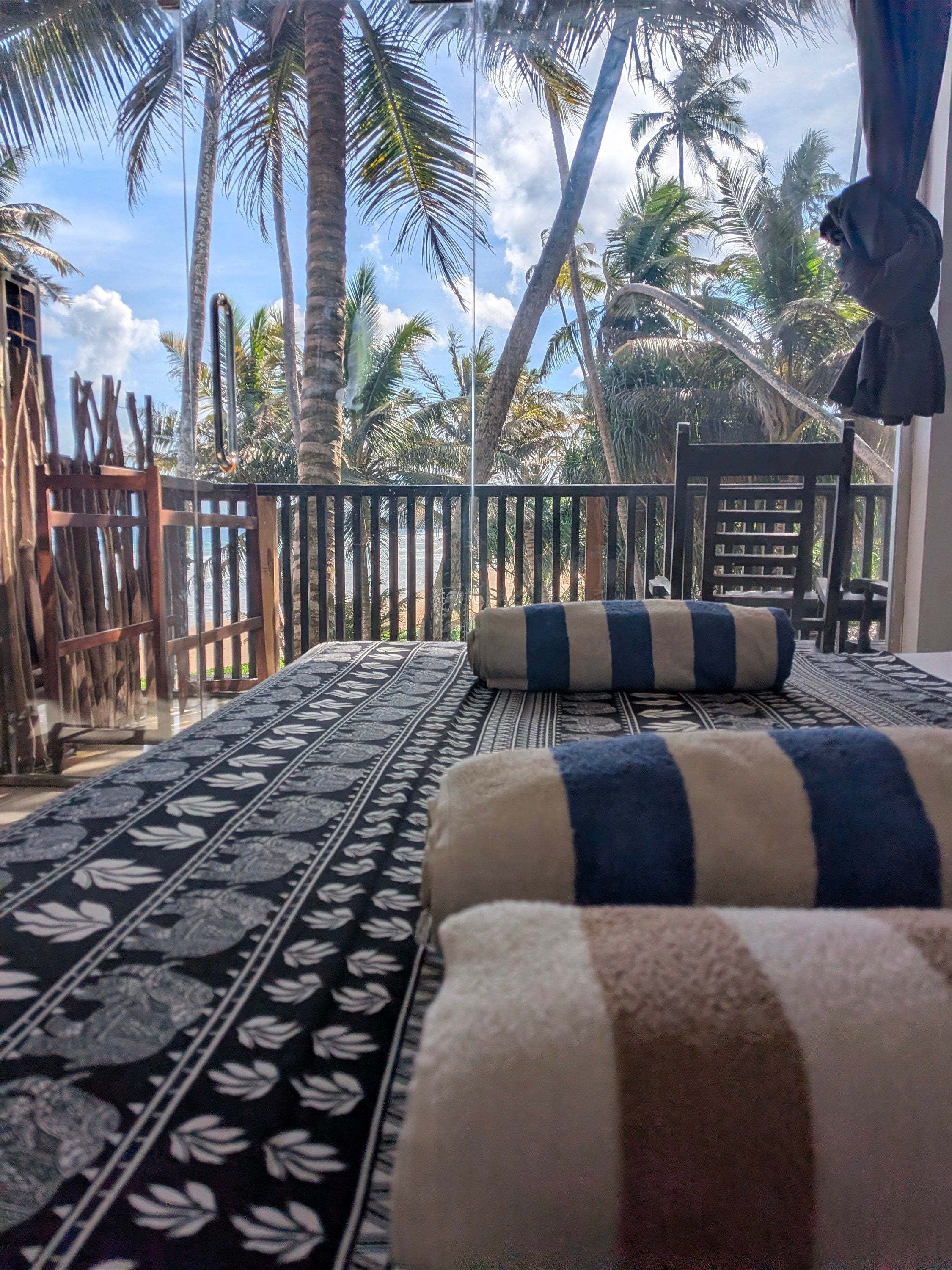 Ein Zimmer mit einem gemusterten Bettüberwurf, einem gestreiften Kopfkissen, einem Holzstuhl und Blick auf Palmen sowie einen Strand.