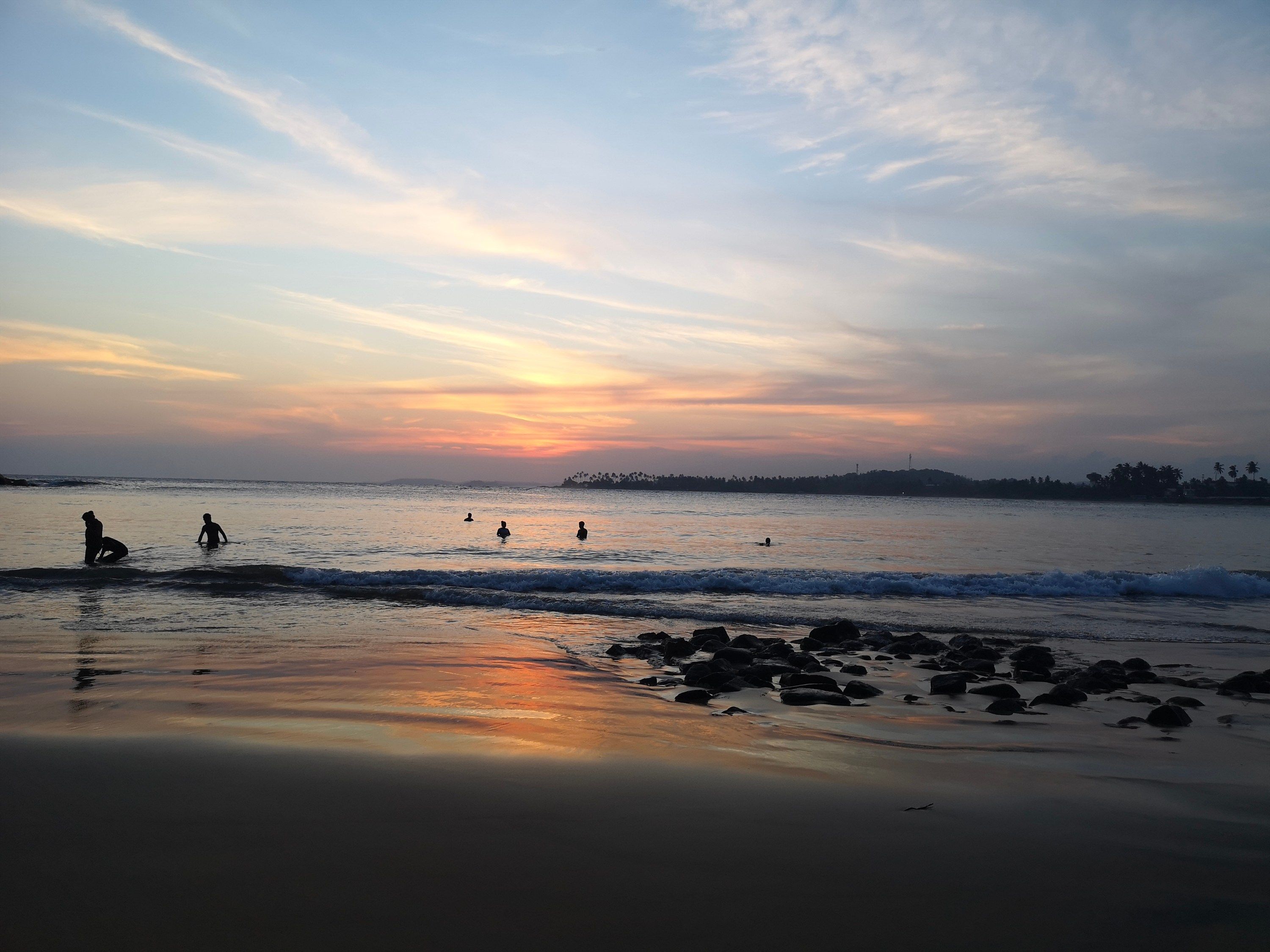 Eine Strandansicht mit Menschen im Wasser und am Ufer, Felsen im Vordergrund und einem Sonnenuntergang im Hintergrund.