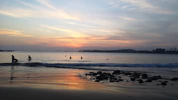 Una spiaggia con persone in acqua e sulla riva, rocce in primo piano e un tramonto sullo sfondo.