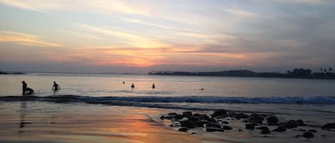 A beach scene with people in the water and on the shore, rocks in the foreground, and a sunset in the background.