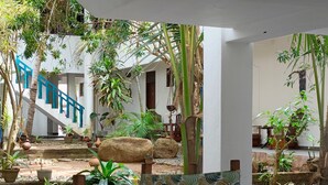 A wooden table with patterned chairs, surrounded by potted plants and a staircase with blue railings leading to a white building. - Dickwella Beach Hotel (Dikwella)
