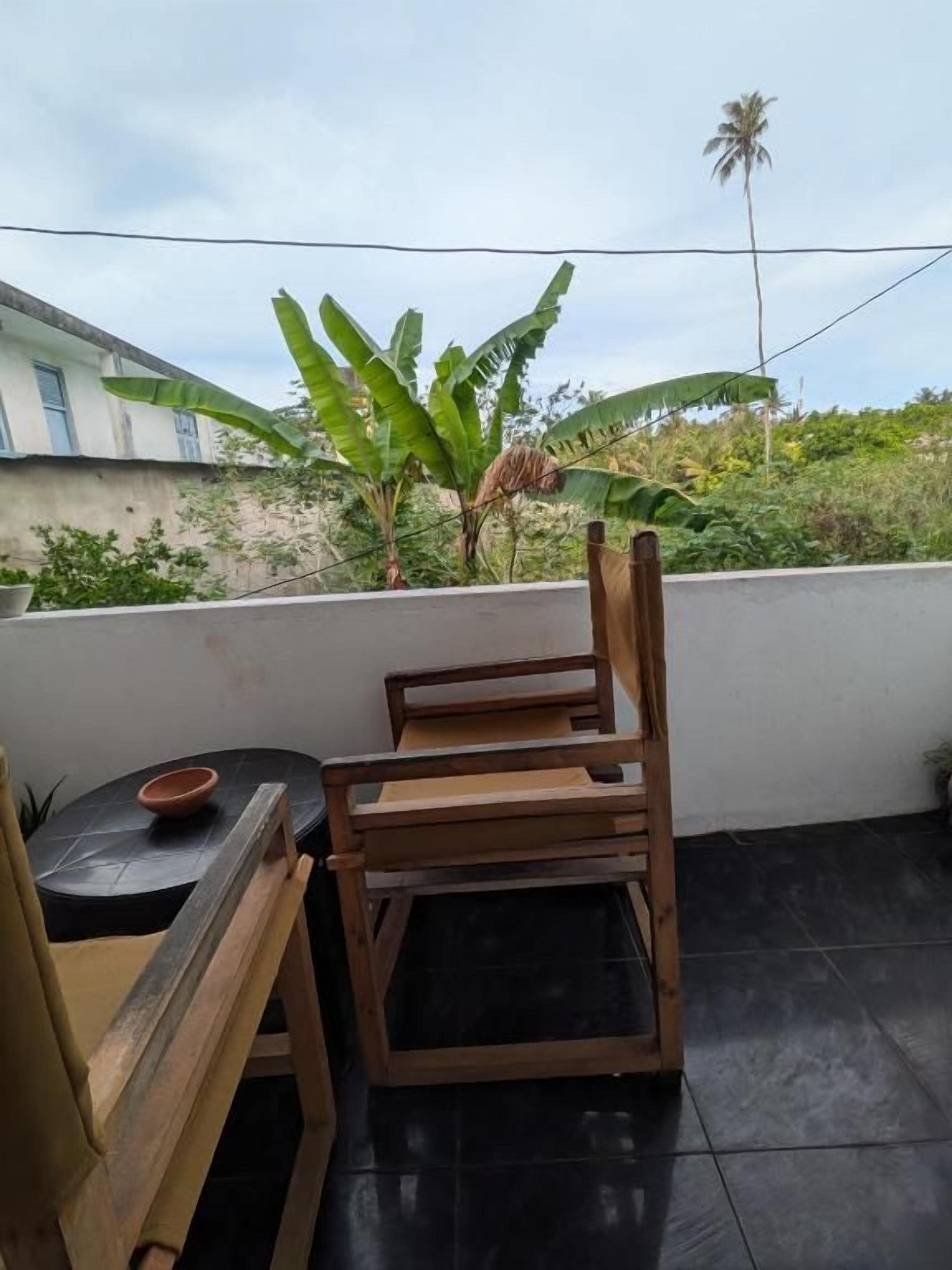 A balcony with two wooden chairs and a small table, a potted plant, and a view of a building and palm tree.