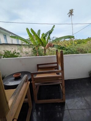 A balcony with two wooden chairs and a small table, a potted plant, and a view of a building and palm tree.