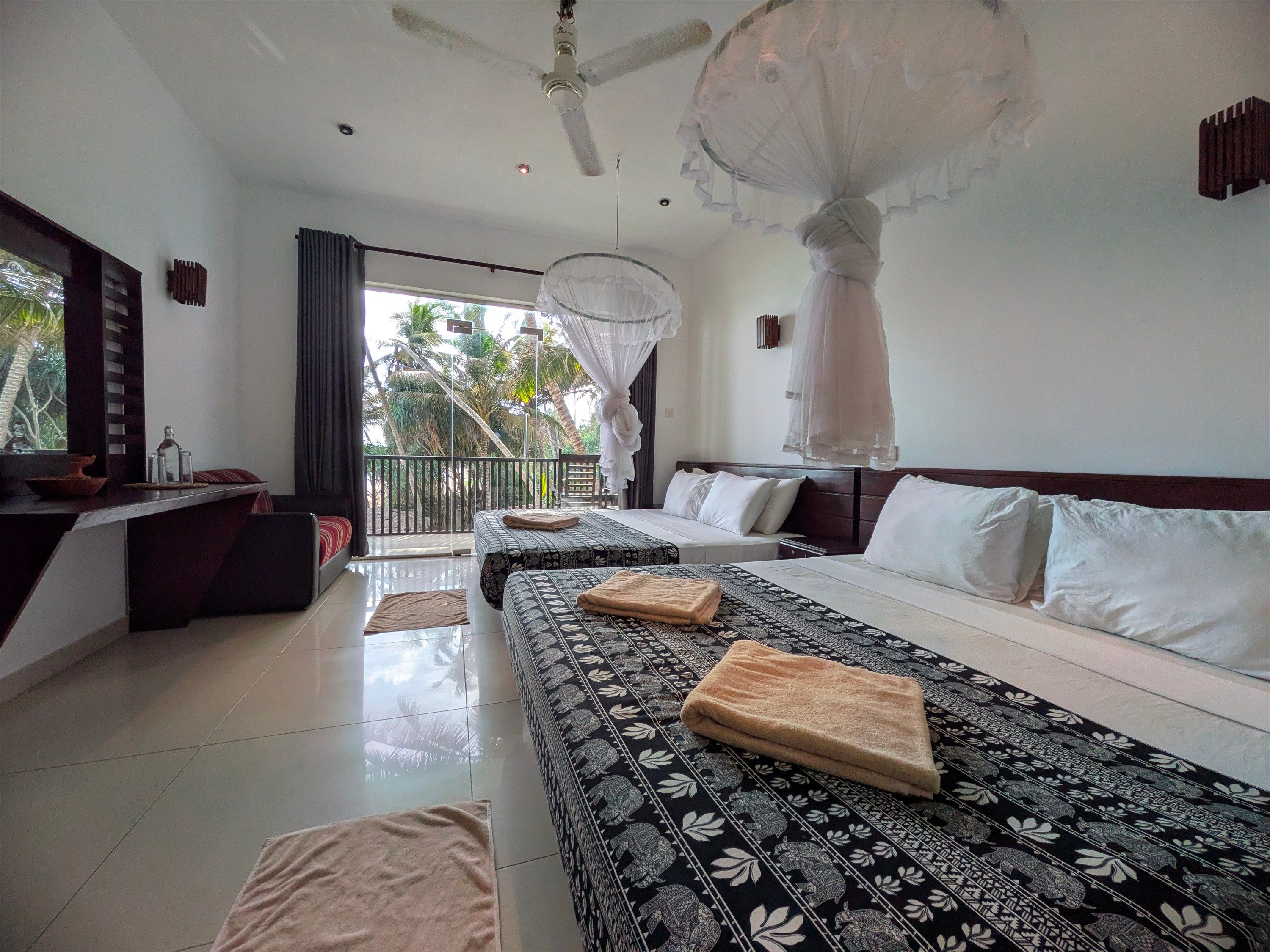 A hotel room with a bed, a sofa, a balcony with a view of palm trees, a ceiling fan, and a mosquito net.