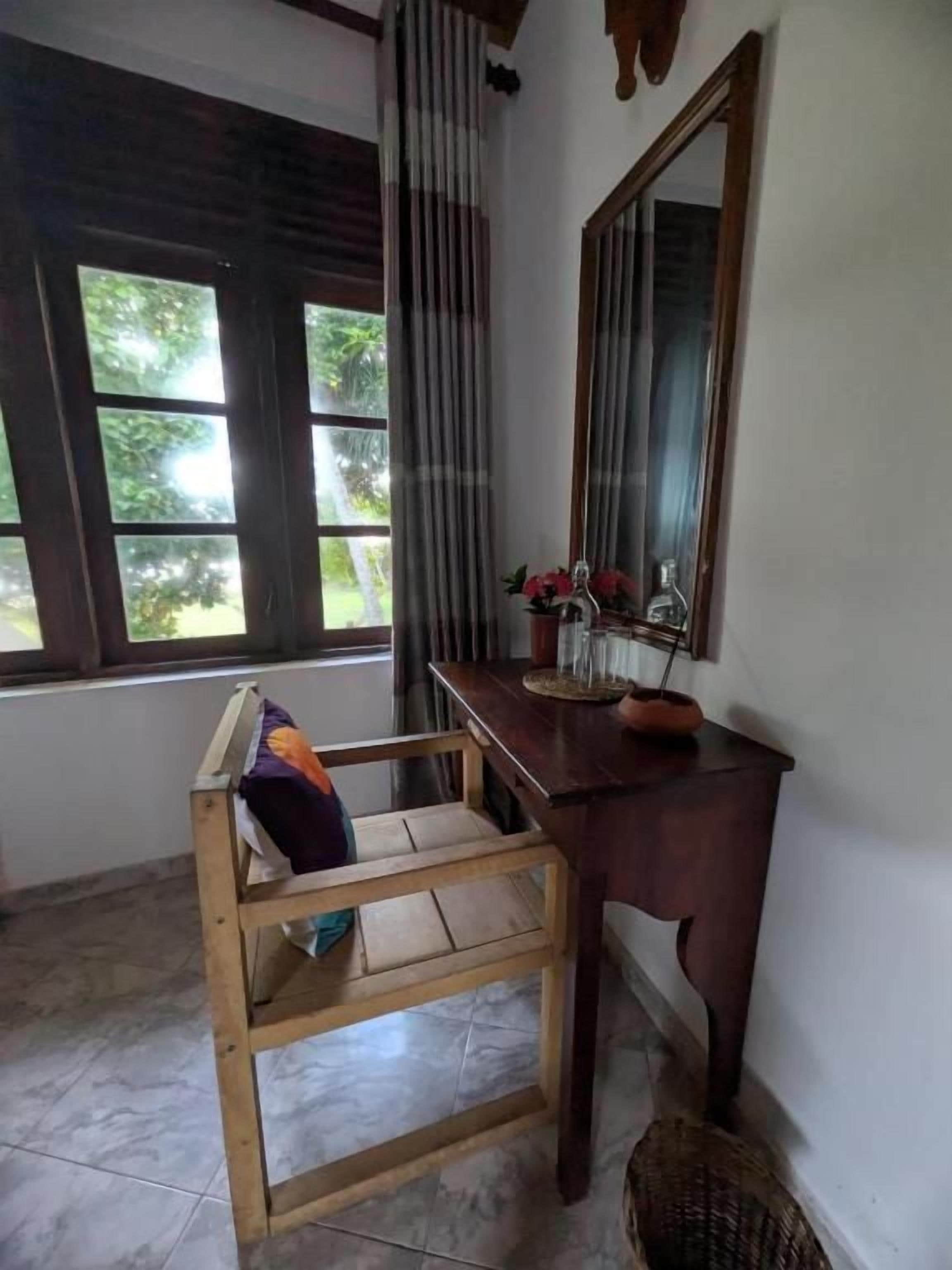 Una habitación con una silla de madera, una mesita con un florero, un espejo y una ventana con cortinas.