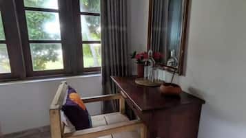 Una habitación con una silla de madera, una mesita con un florero, un espejo y una ventana con cortinas.