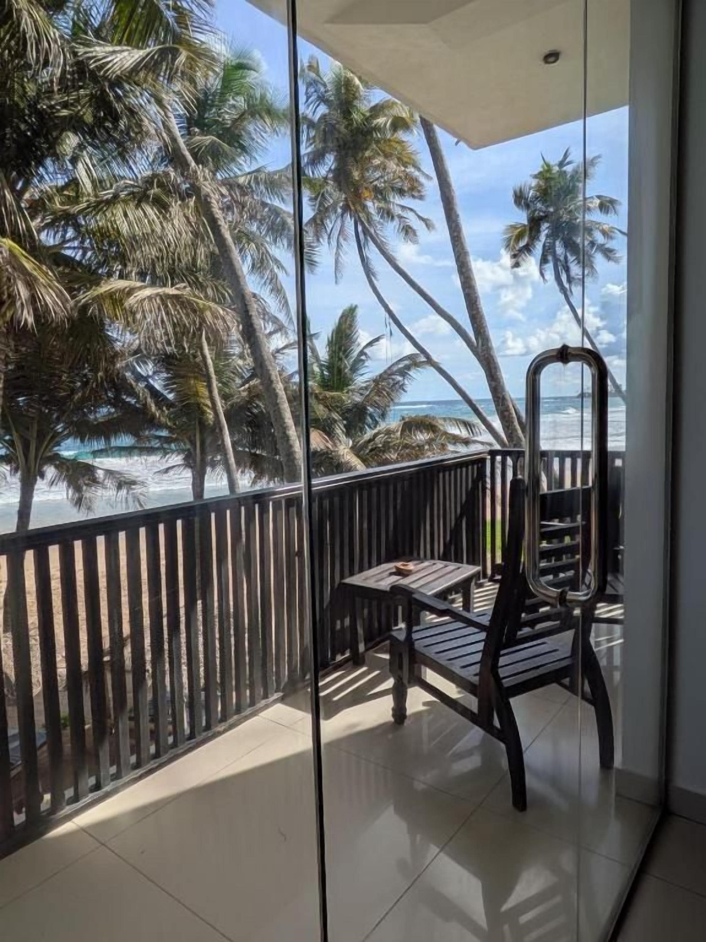 A balcony with a view of the ocean and palm trees, a chair, and a table.
