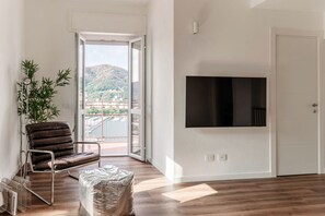 A modern living room with a large flat-screen TV mounted on the wall, a leather sofa, a potted plant, and a sliding glass door leading to a balcony with a view of hills. - Como In The Sky - 150 steps, by MyHomeInComo (Como)