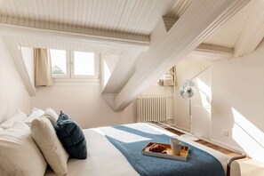 Une chambre à coucher avec un lit, des oreillers, un plateau avec une tasse et une pâtisserie, une lampe de sol et une fenêtre.