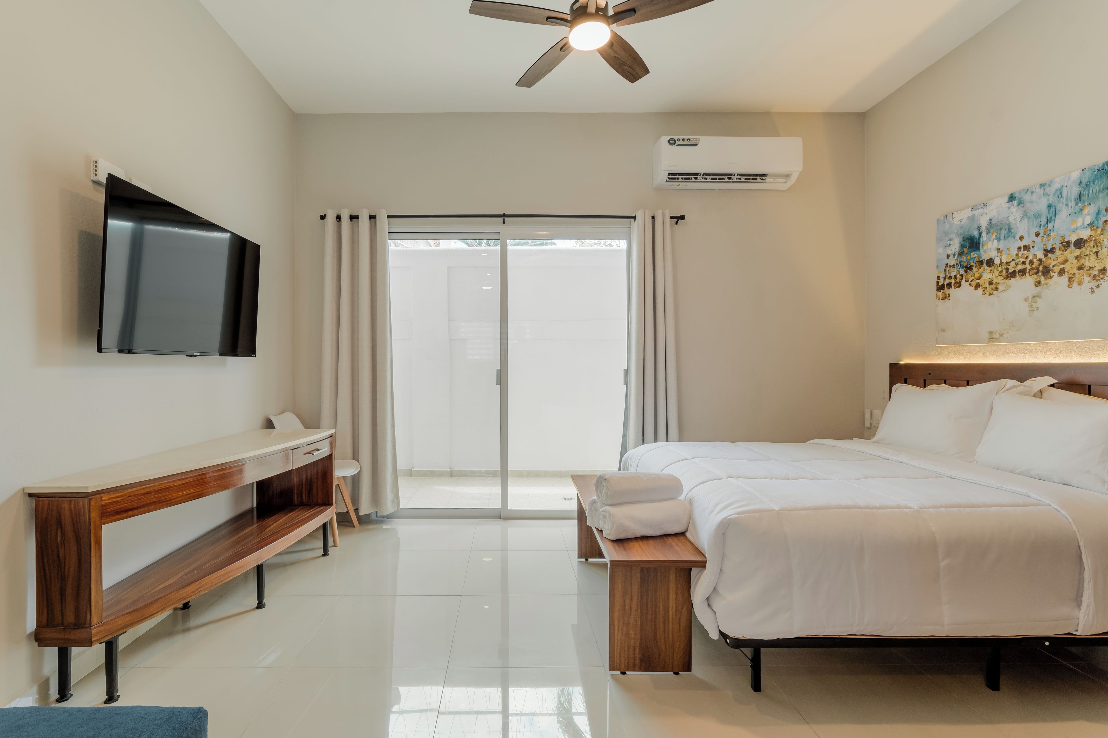 Quarto de hotel com cama, televisão, ventoinha no teto, ar condicionado, pintura e porta de vidro deslizante.