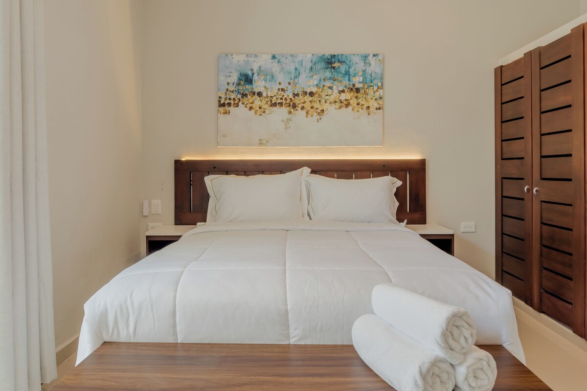 Una habitación de hotel con cama, cabecera, mesita de noche, armario de madera y una obra de arte colgada en la pared.