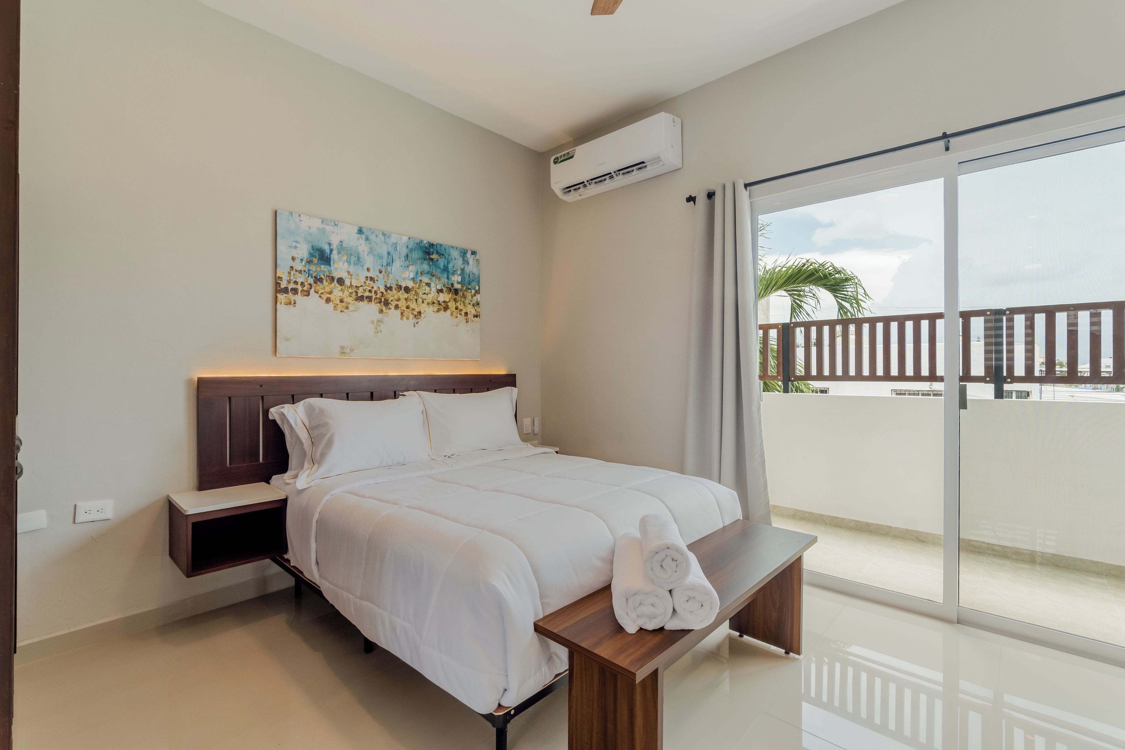 Una camera d'albergo con un letto, una panca, un quadro, un condizionatore d'aria e vista sull'esterno.