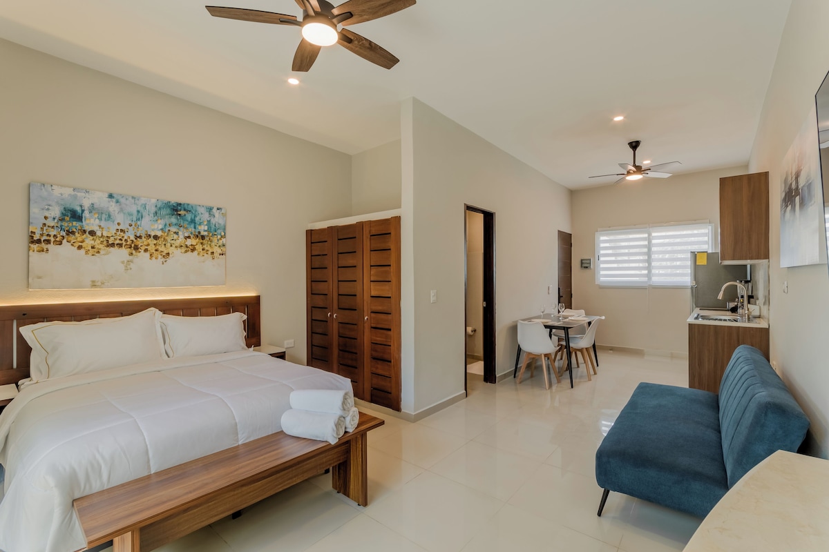 A hotel room with a bed, a desk, a chair, a bench, a ceiling fan, and a painting on the wall.