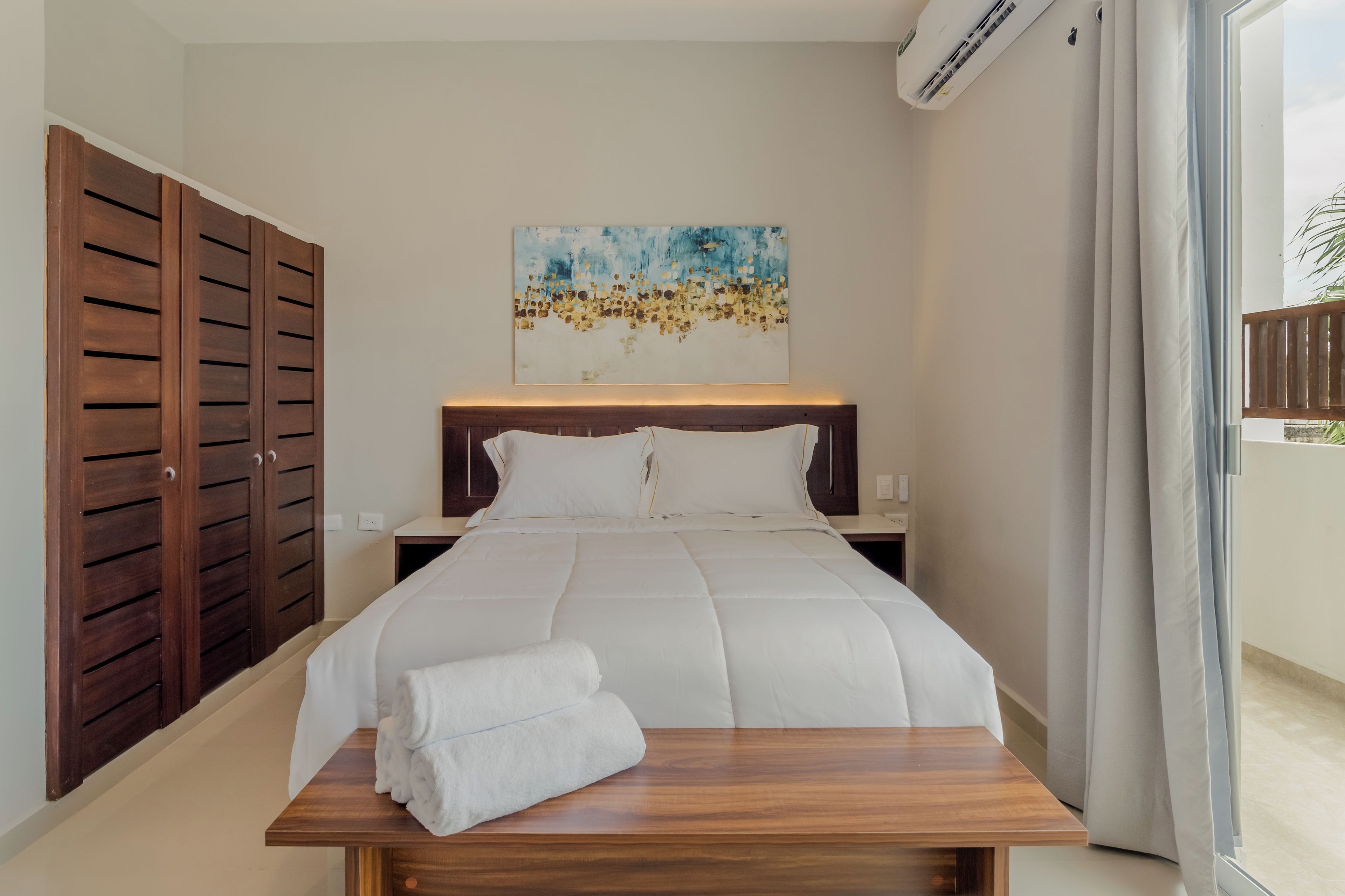 A hotel room with a bed, a wooden nightstand, a painting on the wall, and a view of the outdoors.