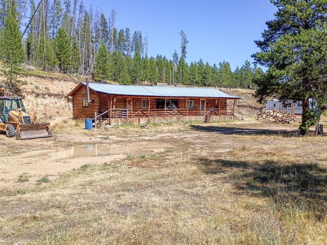 Una cabaña de madera con techo metálico, rodeada por un campo seco y cubierto de hierba y una ladera boscosa.