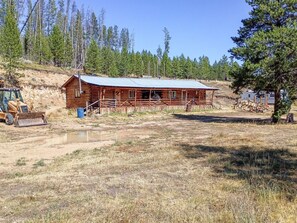 A wooden cabin with a metal roof, surrounded by a dry grassy field and a forested hillside.