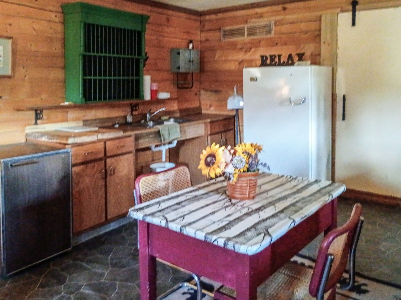 Interior de uma cabana de madeira com cozinha, mesa de jantar e um vaso de flores, além de uma geladeira com a etiqueta 'RELAX'.