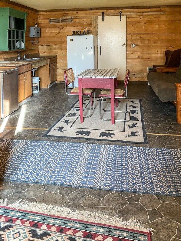 A rustic cabin interior with a wooden dining table, chairs, a refrigerator, and a kitchen area.