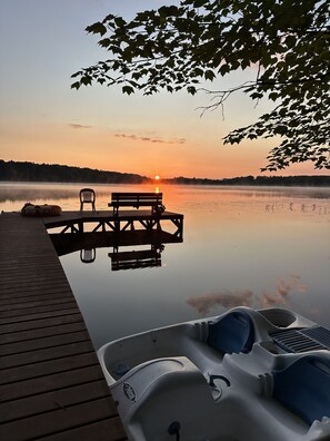 Ein stiller See bei Sonnenuntergang mit einem Steg, einem Boot und einer Bank.