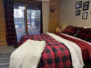 A room with a bed, a window with curtains, a wall with framed pictures, and a wooden floor. - Birch Tree Retreat - Cozy 2BR Lake Escape on Eco Lake (Eagle River)