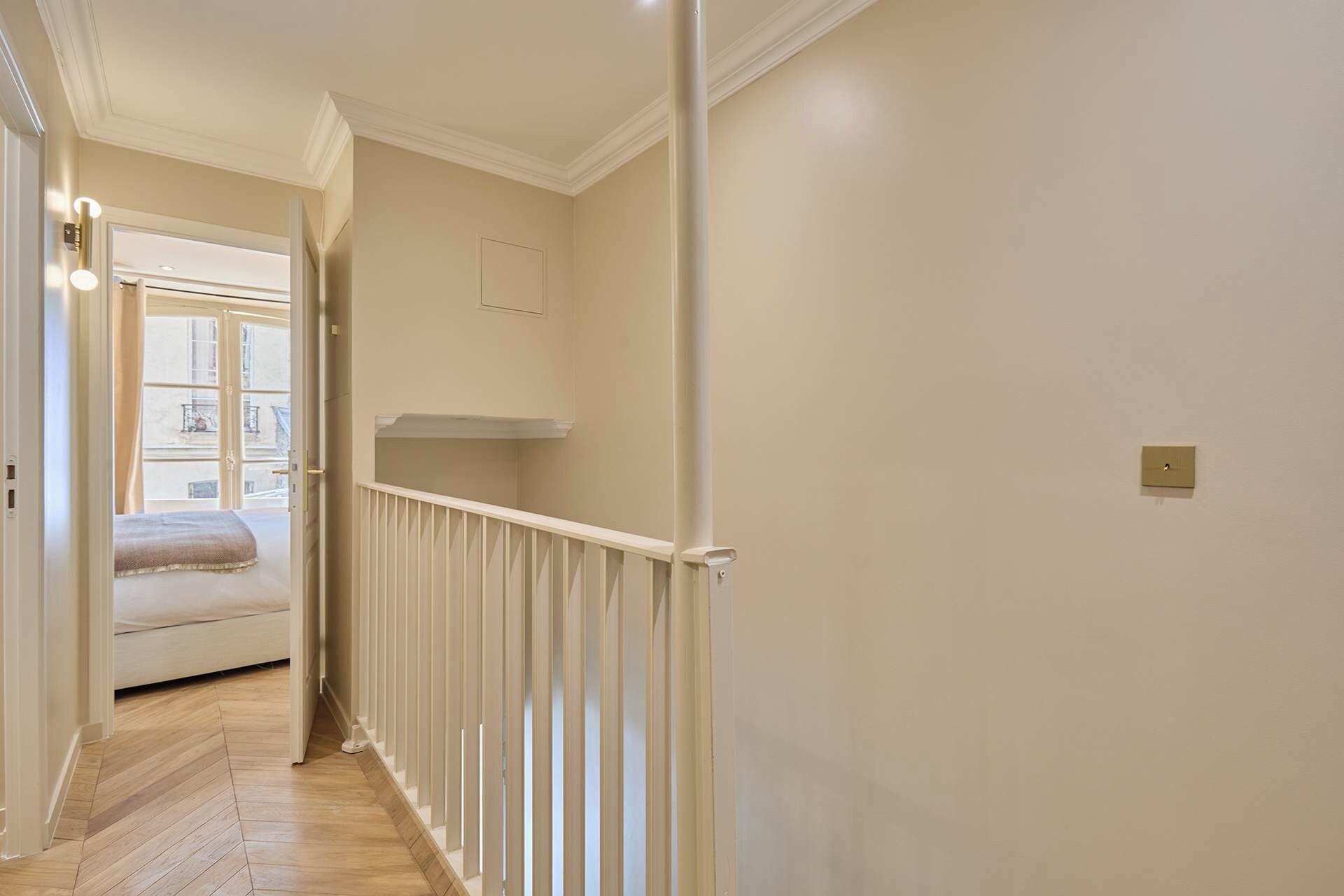 Un pasillo estrecho con barandilla blanca, una cama al final y una ventana con vistas a edificios.