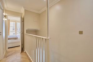 Un couloir étroit avec une rambarde blanche, un lit au fond et une fenêtre donnant sur des immeubles.