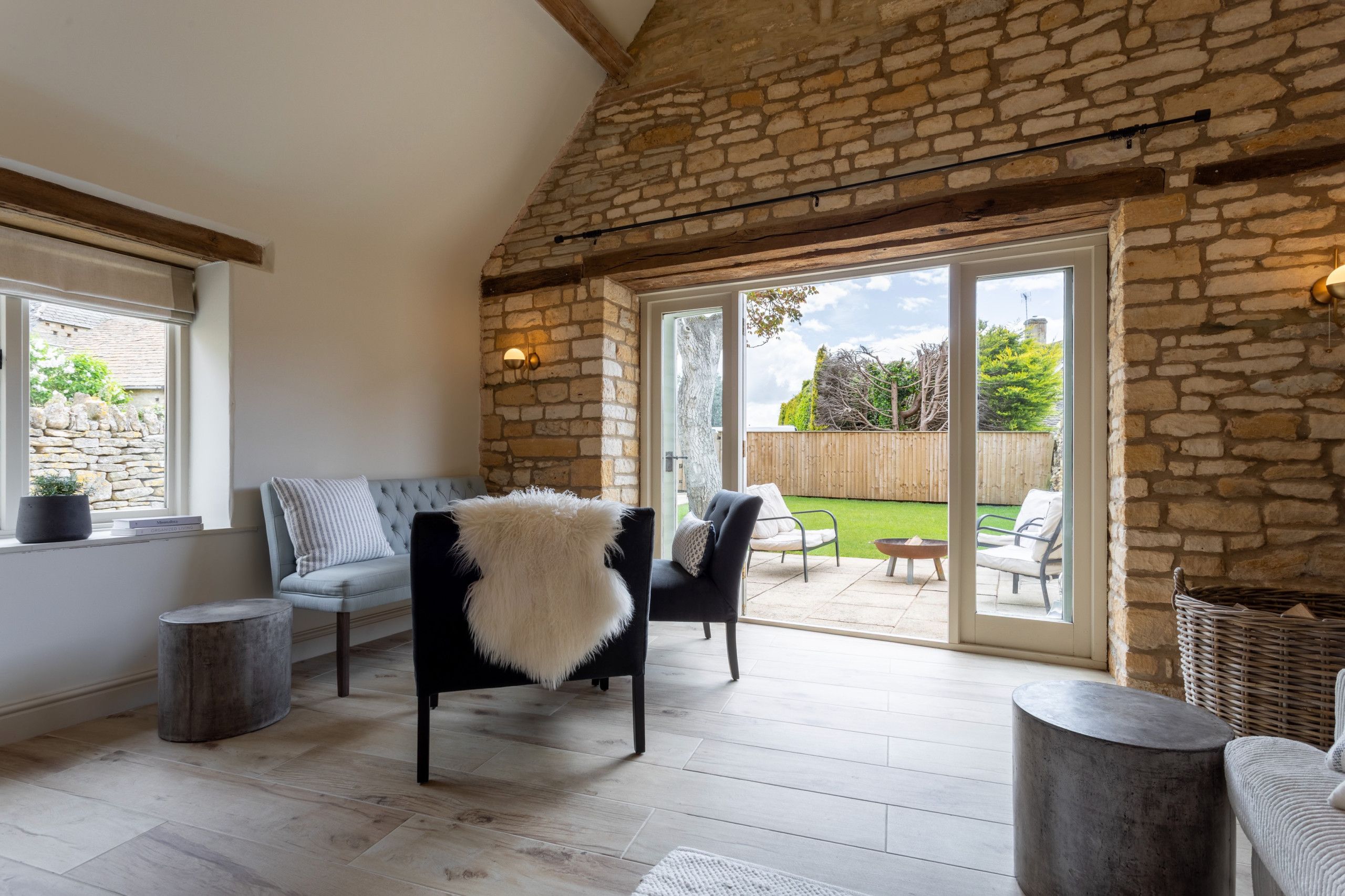 A room with a stone wall, a black chair with a white throw, a grey sofa, and a view of a garden through a glass door.