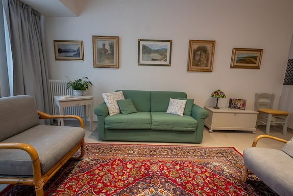 Un salon avec un canapé vert, deux fauteuils, un tapis à motifs et quatre tableaux encadrés accrochés au mur.