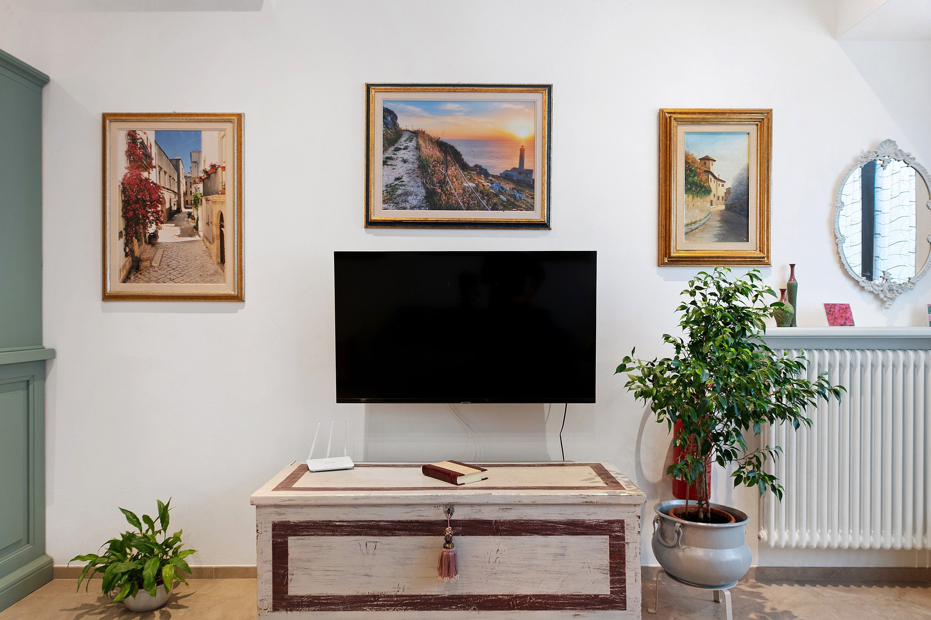 A living room with a flat-screen TV, three framed paintings, a mirror, and a potted plant.