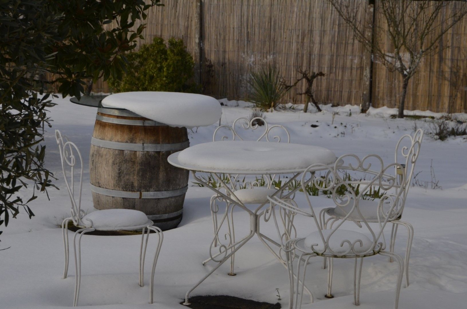 Een terras in de open lucht, bedekt met sneeuw, met een metalen tafel en stoelen, een houten vat en een bamboe hek op de achtergrond.