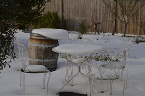 Ein mit Schnee bedeckter Außenbereich mit einem Metalltisch und -stühlen, einem Holzbottich und einem Bambuszaun im Hintergrund.