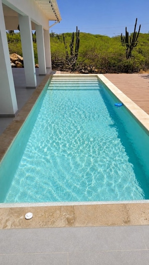 A rectangular swimming pool with clear blue water, surrounded by a wooden deck and a white structure with columns. - Cas Seida - Private Pool Villa Pure Aruba (Oranjestad)