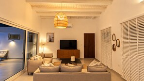 A modern living room with a sofa set, a flat-screen TV, a wooden cabinet, and a large window with blinds. - Cas Seida - Private Pool Villa Pure Aruba (Oranjestad)
