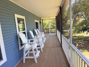A covered porch with white Adirondack chairs, a wooden deck, and a view of a grassy area with trees. - Heaven on Earth- Intracoastal Waterfront, 15 minutes from Emerald Isle (Hubert)