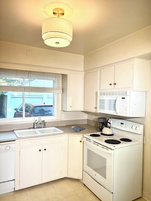 A kitchen with white cabinets, a microwave, an oven, a sink, and a window with blinds.