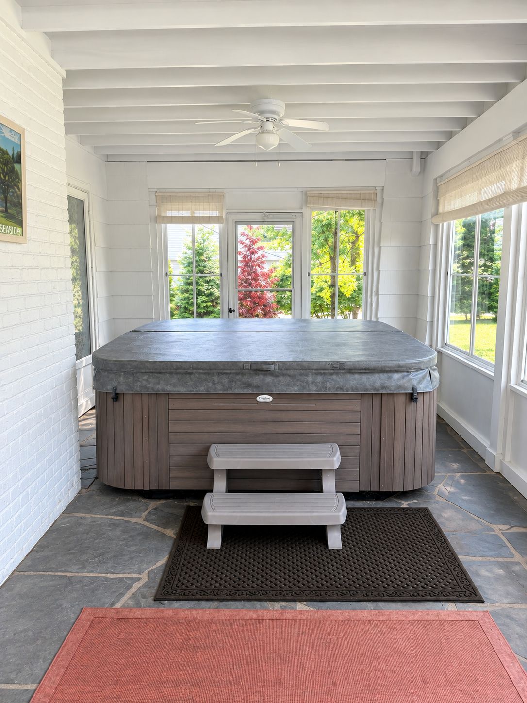 A hot tub with a wooden bench in a room with white walls, large windows, and a ceiling fan.
