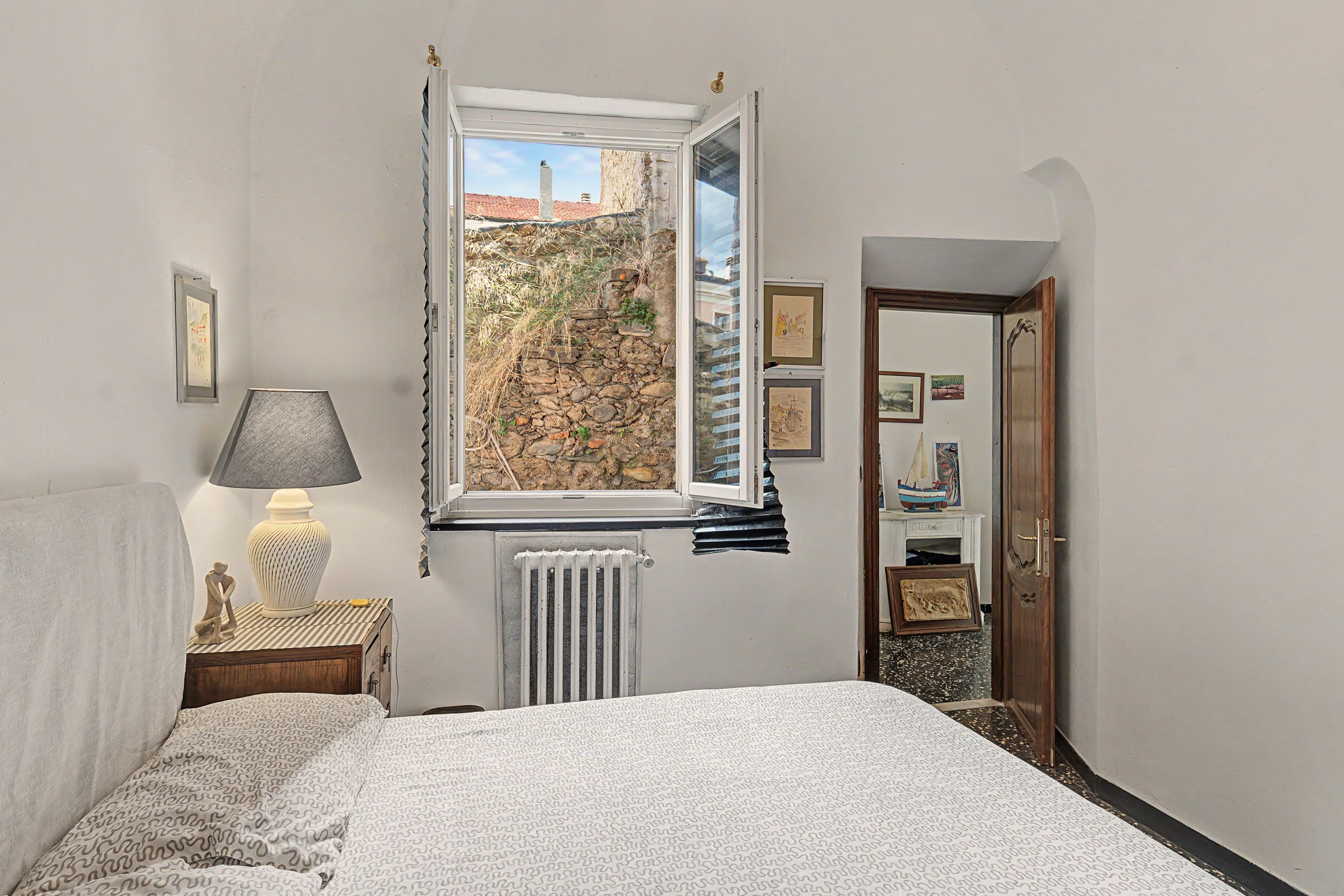 Ein Schlafzimmer mit einem Bett, einem Nachttisch mit Lampe, einem Heizkörper, einem Fenster mit Blick auf ein Gebäude und einer Tür.