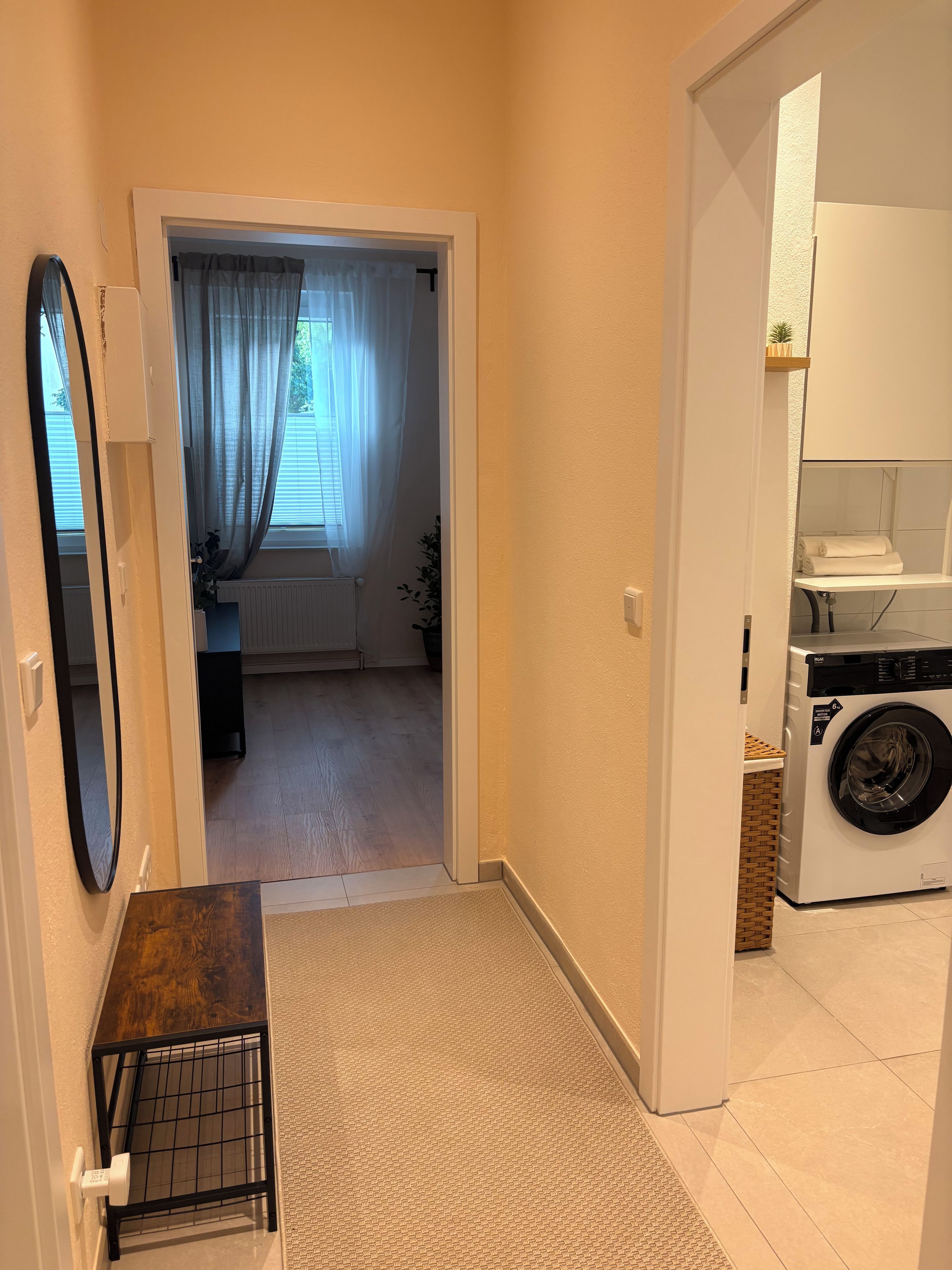 A hallway leading to a laundry area with a washing machine, a mirror, and a door to a room with a radiator and curtains.
