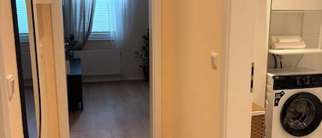 A hallway leading to a laundry area with a washing machine, a mirror, and a door to a room with a radiator and curtains.