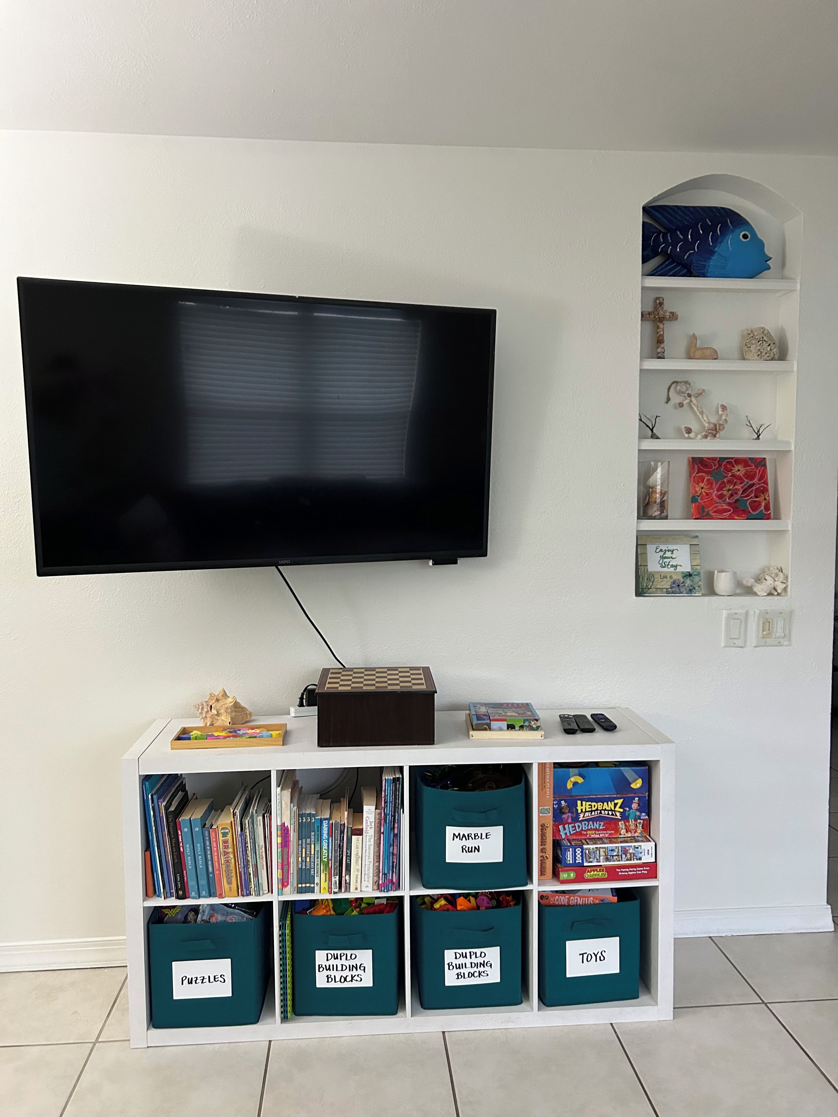 A living room with a flat-screen TV mounted on the wall, a white entertainment unit below with various storage bins labeled for toys, puzzles, and building blocks, and a shelf with decorative items and a book.