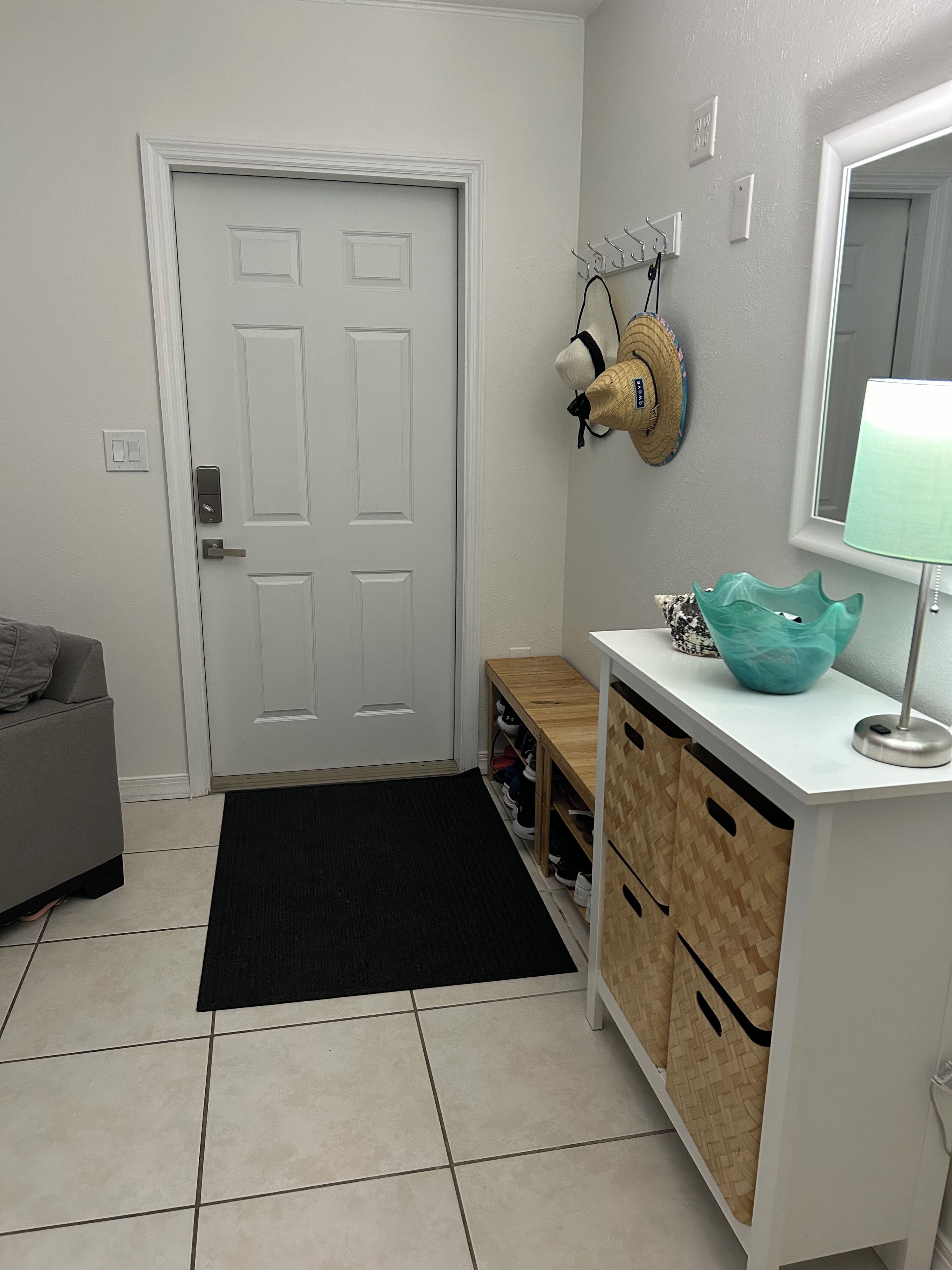 A hallway with a white door, a bench, a shoe rack, a mirror, and a teal bowl on a white counter.