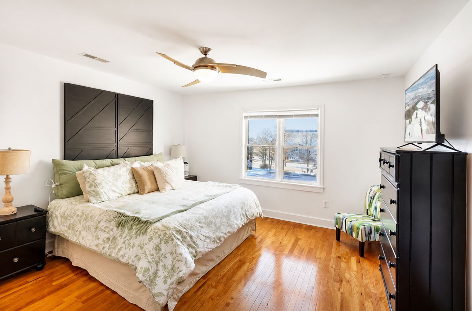 A bedroom with a bed, nightstands, a dresser, a ceiling fan, a window with a view, and a dresser with drawers.