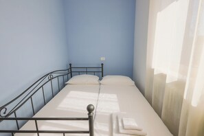 A single bed with a metal frame, white bedding, and a window with curtains on the right.