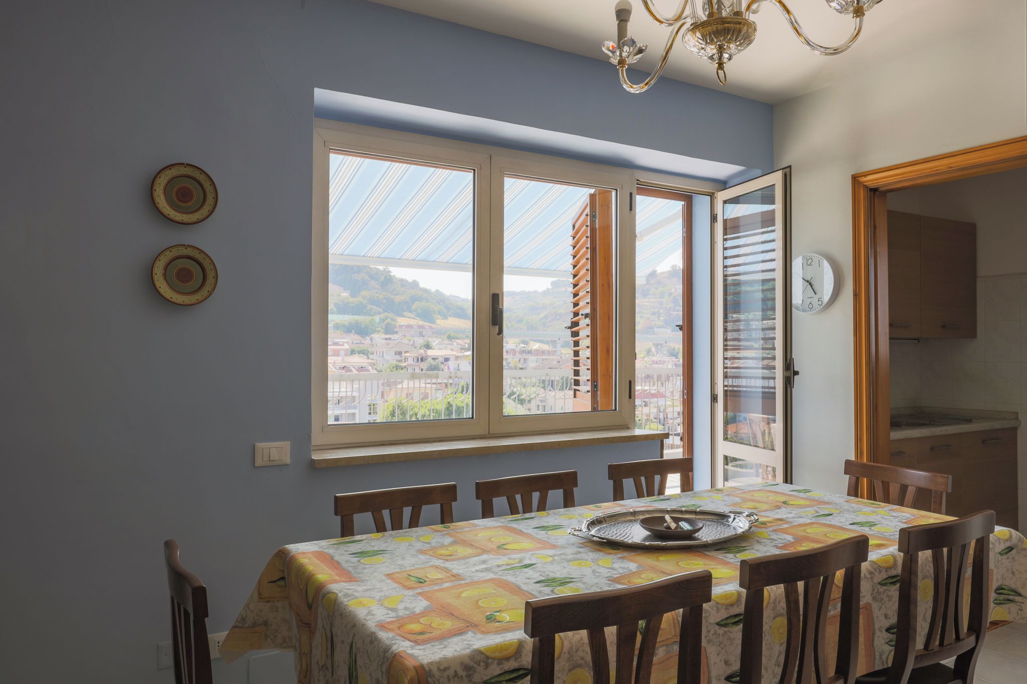 A dining area with a table set for six, a view of a town through a window, and a kitchen visible through an open door.