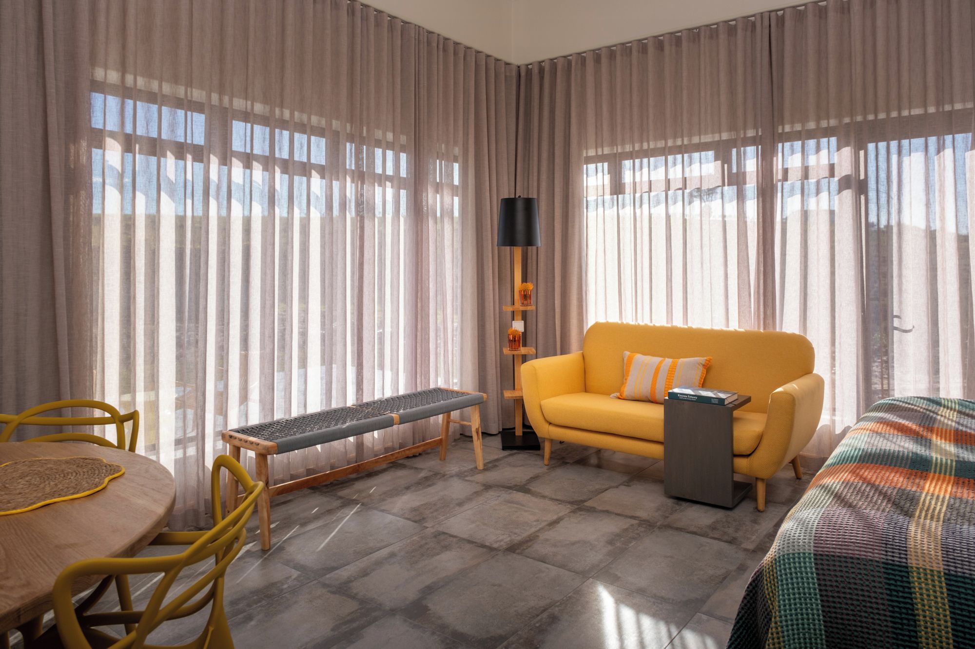 A hotel room with a yellow sofa, a small table, a bench, and a bed with a colorful blanket.
