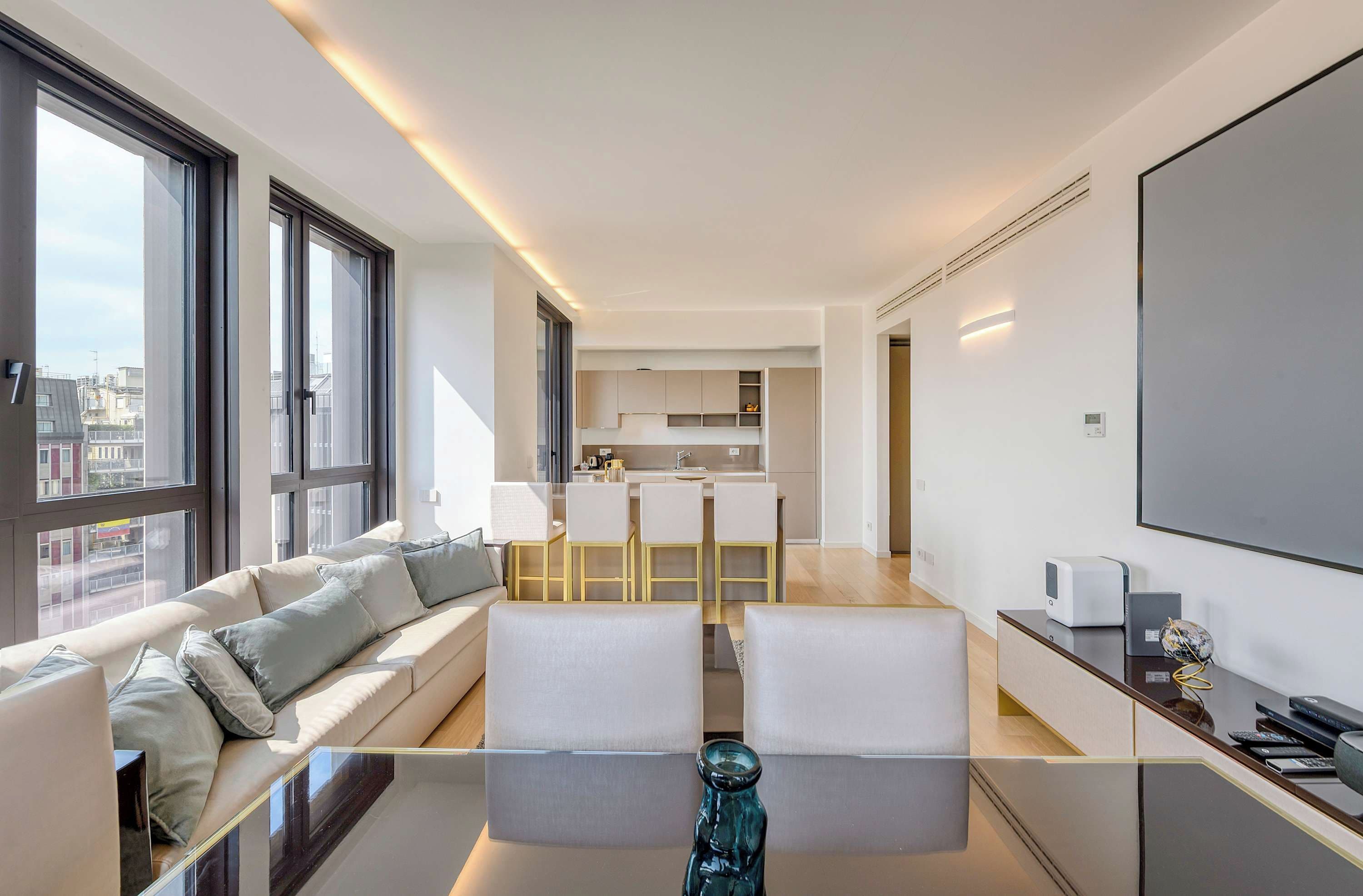 A modern apartment with a living area featuring a sofa and a glass coffee table, a kitchen with a dining area, and large windows offering a city view.
