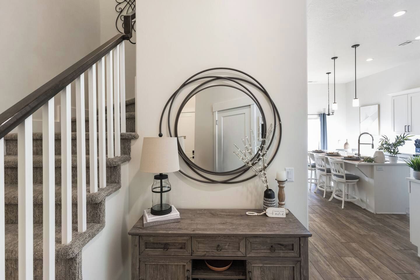A staircase with a wooden handrail and carpeted steps, a wooden console table with a lamp, a mirror, and decorative items, and a kitchen area with bar stools and a central island.