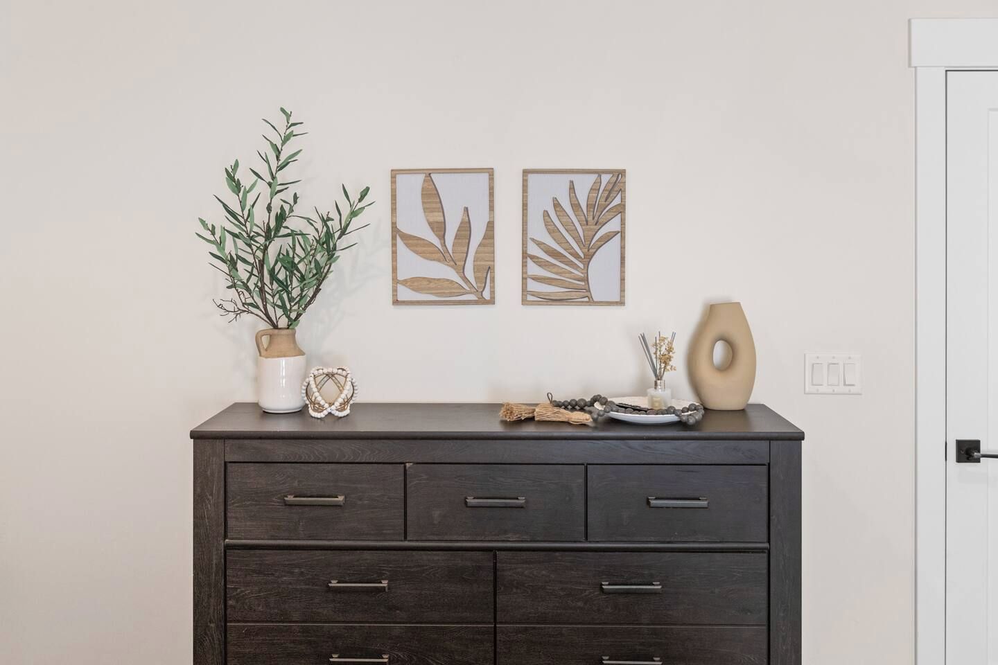 Une commode en bois foncé, dotée de nombreux tiroirs, une plante en pot et des objets décoratifs, notamment un vase, un collier de perles et deux œuvres d’art représentant des feuilles, encadrées.