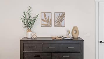 Eine dunkle Holzkommode mit mehreren Schubladen, eine Topfpflanze und Dekorationselemente wie eine Vase, eine perlenbesetzte Halskette und zwei gerahmte Blätter-Kunstwerke.