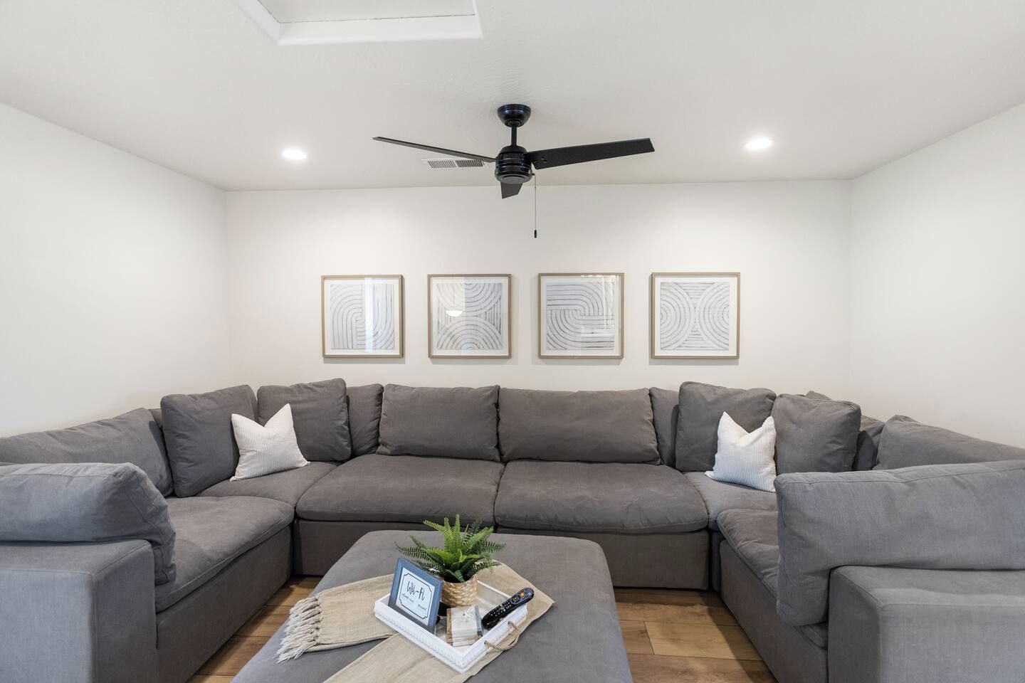A modern living room with a grey sectional sofa, a ceiling fan, and four framed abstract wall art pieces.
